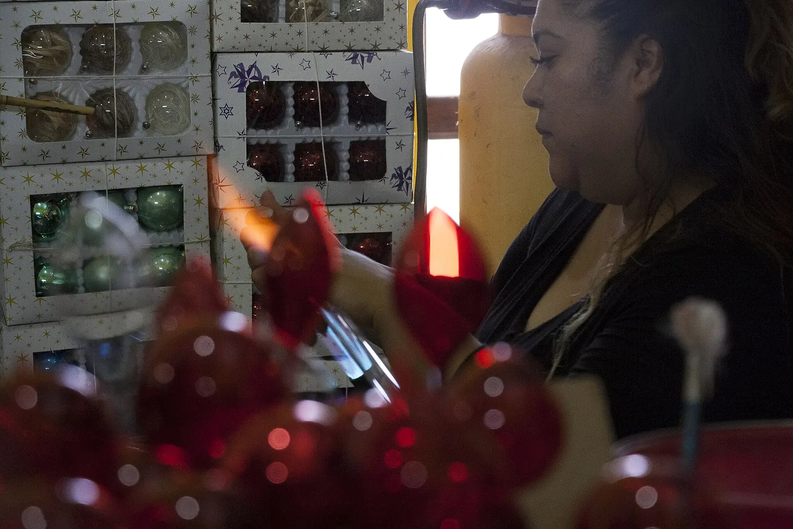 Artesana soplando una esfera de vidrio en un taller de Chignahuapan, Puebla, con cajas de esferas navideñas al fondo
