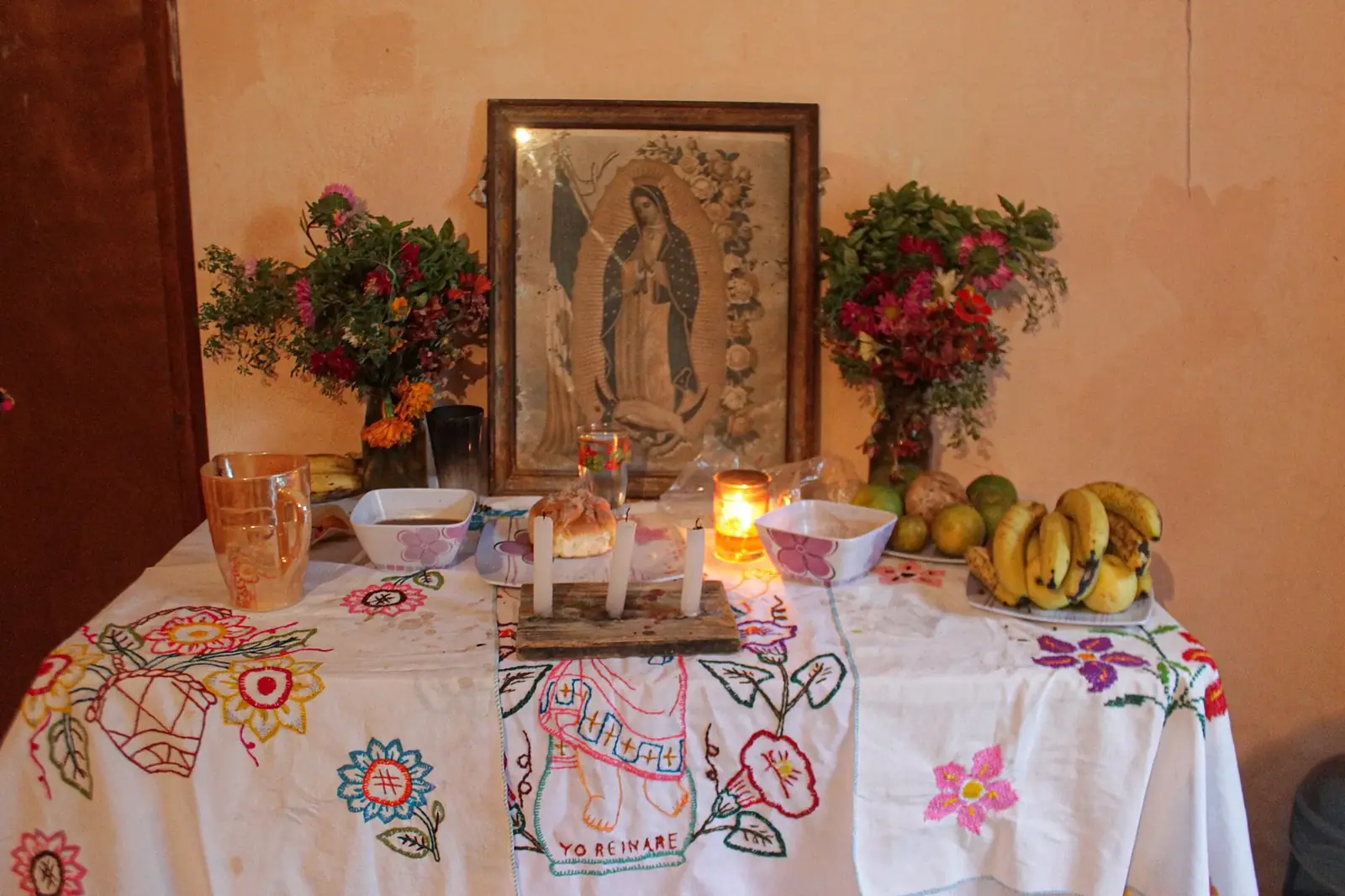 Altar de Hanal Pixán en una casa de Yucatán: mantel bordado, velas, pan, fruta y flores ante una imagen de la Virgen de Guadalupe